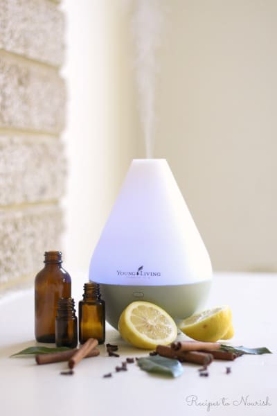 Young Living essential oil diffuser with amber glass essential oil bottles, cinnamon sticks, cloves, herbs and fresh lemon halves.