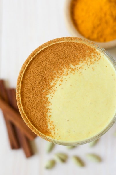 Glass of golden milk smoothie with ground cinnamon dusted on the top and a bowl of ground turmeric with cinnamon sticks.