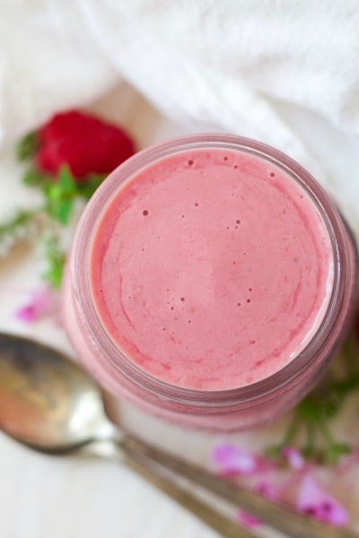 Overhead shot of a jar with pink raspberry smoothie next to fresh raspberries, flowers and a spoon.