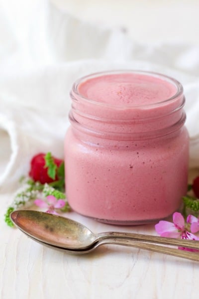 Jar of pink raspberry smoothie next to fresh raspberries, flowers and spoons.
