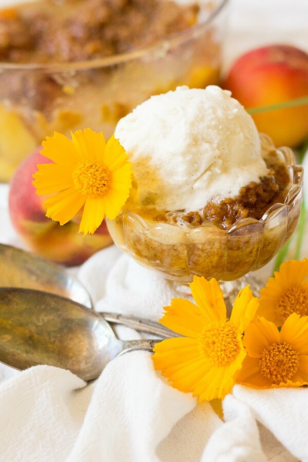 Scoop of vanilla ice cream on top of peach crumble with fresh peaches and orange flowers.