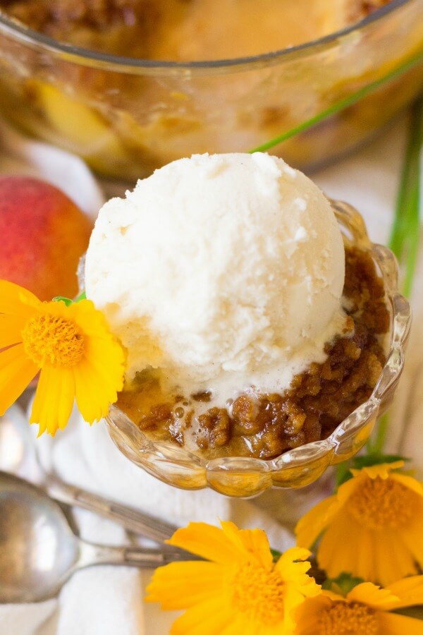 Scoop of vanilla ice cream on top of peach crumble with fresh peaches and orange flowers.