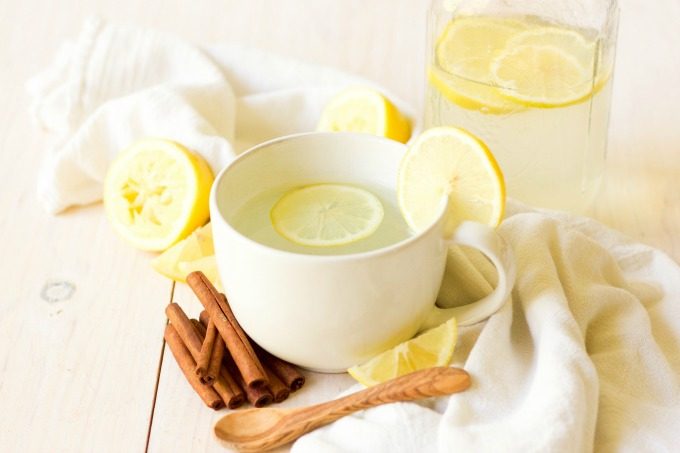 Mug and mason jar full of hot drink and lemon slices next to lemons and cinnamon sticks.